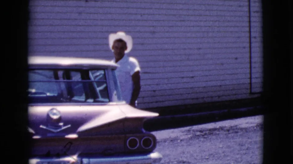 In 1964 Cottonwood, Arizona footage, a young man in a white cowboy hat stands near the rear of a classic car against horizontal siding.