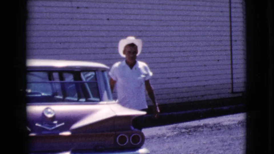 The man faces forward beside the car's rear quarter, with horizontal siding and sunlit gravel visible in the 1964 Cottonwood home movie.