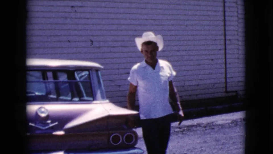 The man walks away from the car's rear area, wearing a white cowboy hat and light shirt in the Cottonwood, Arizona 1964 footage.
