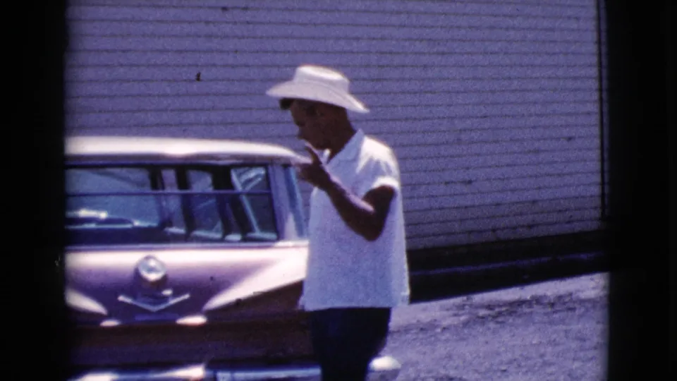 In profile, the man raises a hand near his face while standing beside the tailfin car in the 1964 Cottonwood home-movie shot.
