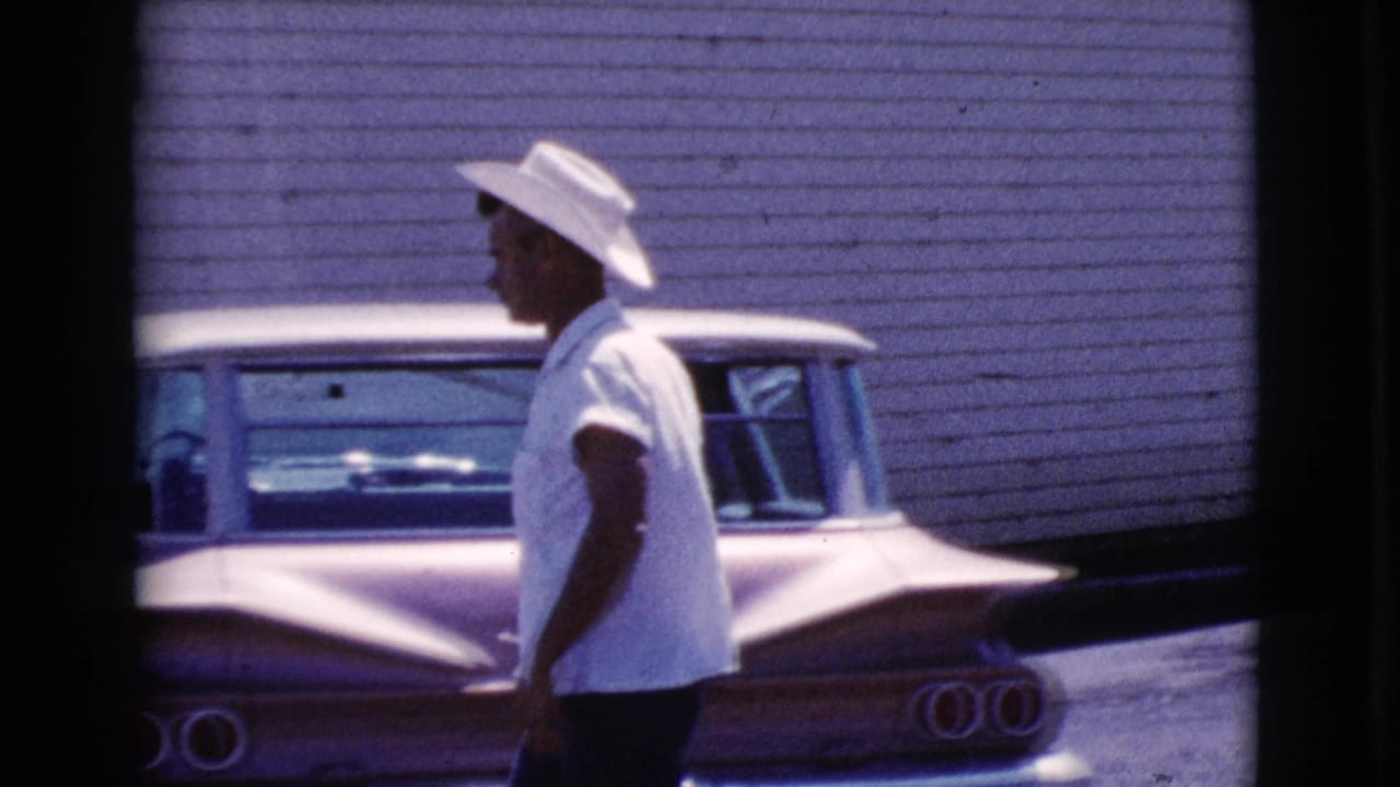 Man in a white cowboy hat walks past a two-tone classic car beside a clapboard building.