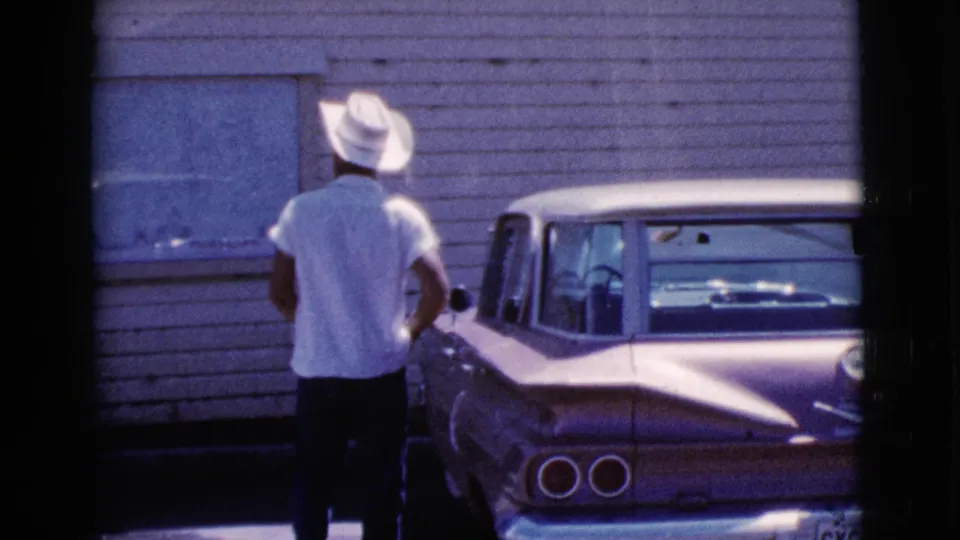 Seen from behind, the man in a white hat stands close to the parked classic sedan near a windowed clapboard wall in 1964 Cottonwood.