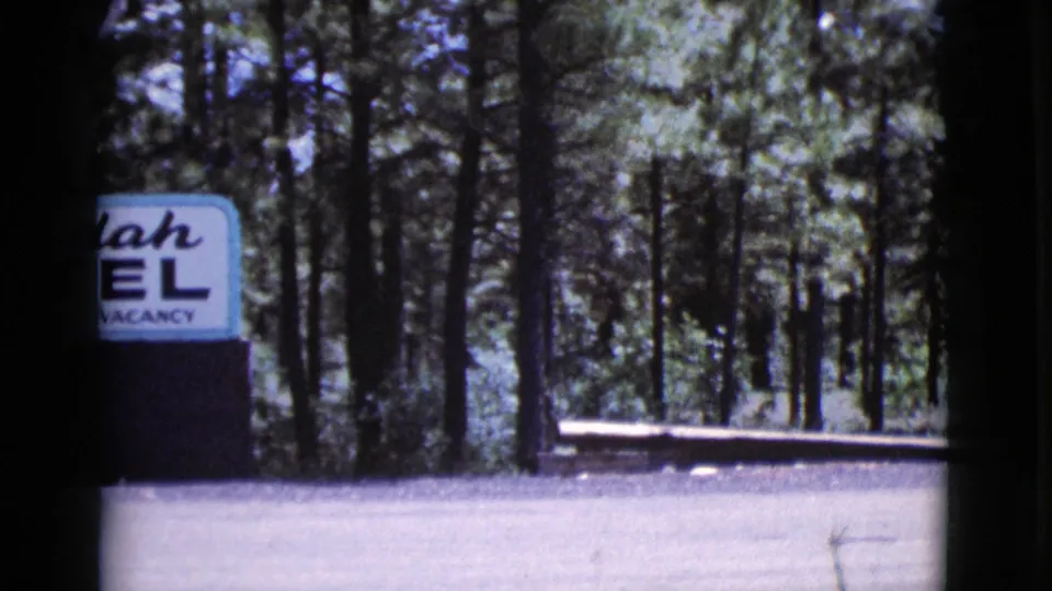 A partly cropped motel sign appears beside a wooded roadside area in the 1964 Cottonwood, Arizona travel footage.