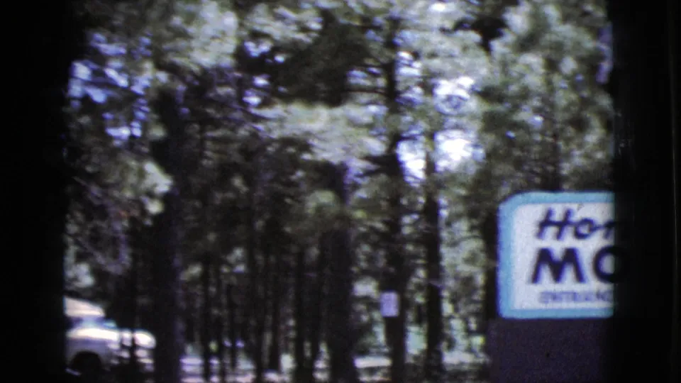 1964 Cottonwood, Arizona 8mm frame showing a partly visible Hon-dah Motel sign among pine trees and a white vehicle near the road.
