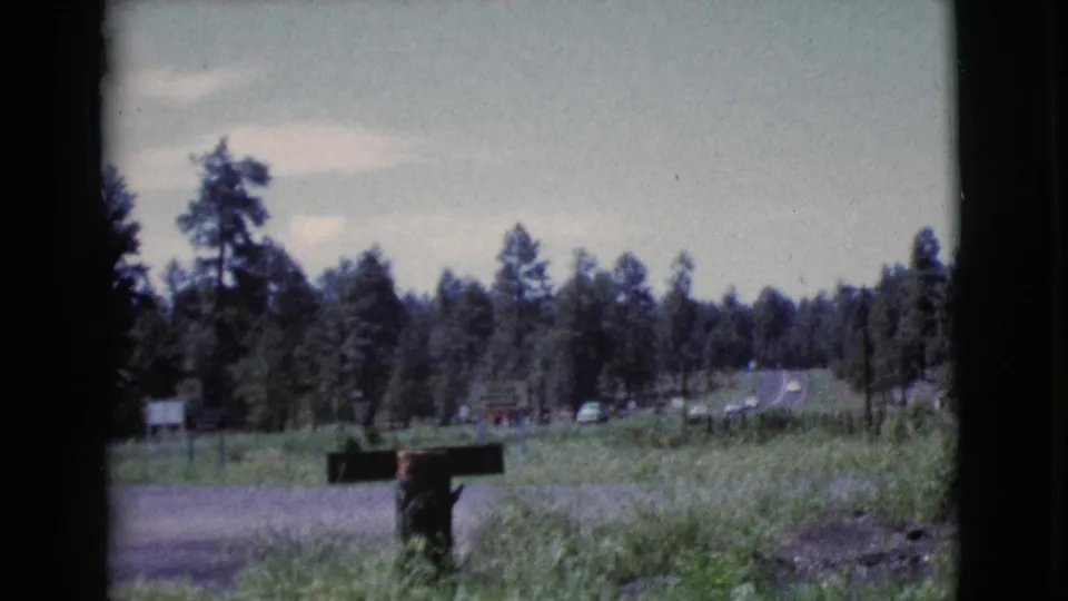 1964 Cottonwood, Arizona 8mm frame with a roadside mailbox, pine trees, grassy shoulder, and distant signage near the motel area.