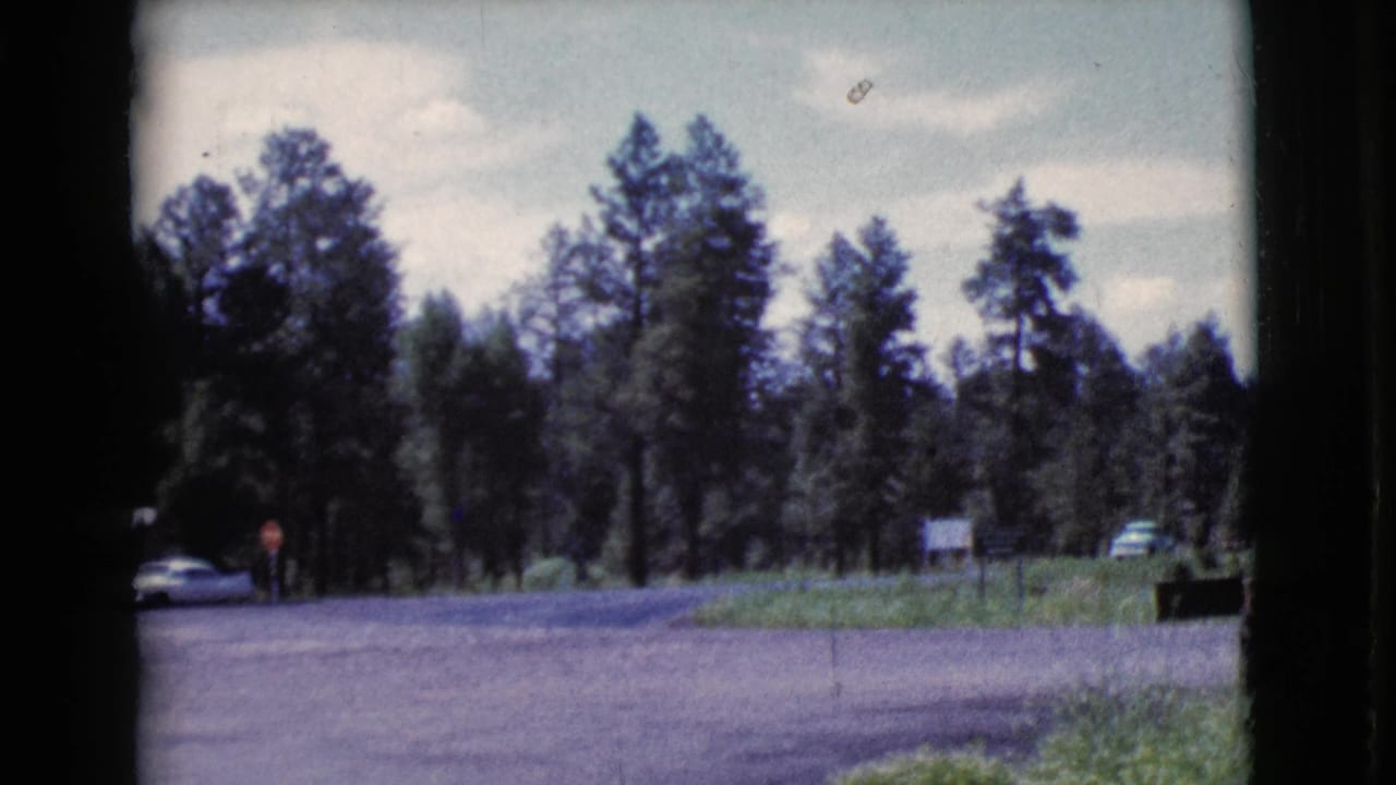 A Cottonwood, Arizona roadside view shows a car at the left edge, distant people, trees, and a signpost in a 1964 8mm frame.