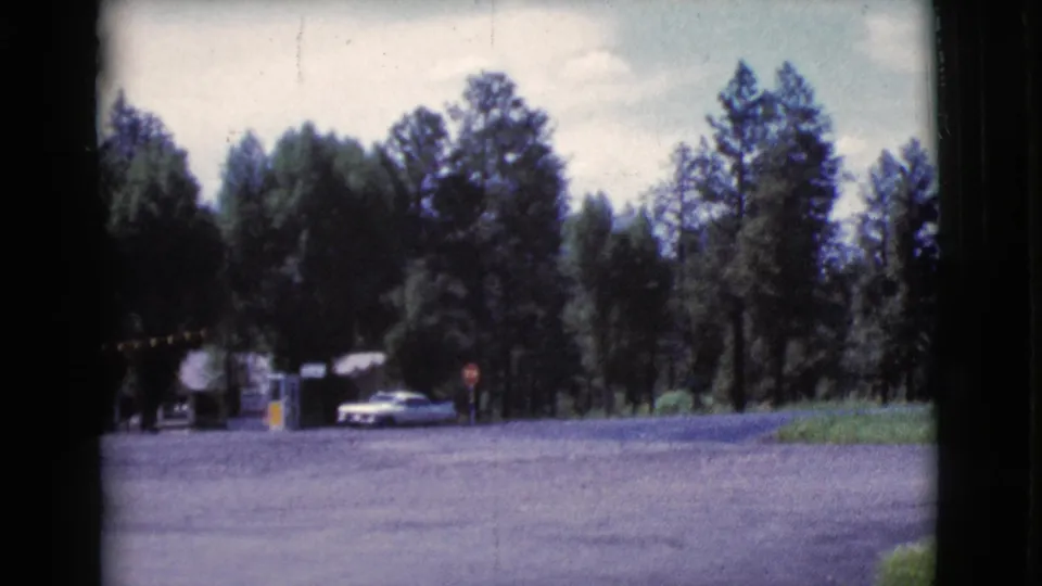 A 1964 Cottonwood, Arizona travel frame shows cars near a small roadside shelter, with distant people standing by a wooded gravel lot.