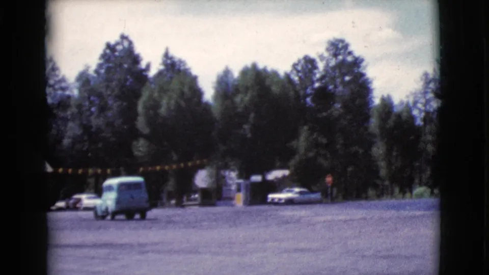 A turquoise wagon or van moves across a wooded gravel roadside area in Cottonwood, Arizona, with other cars and visitors in the distance.