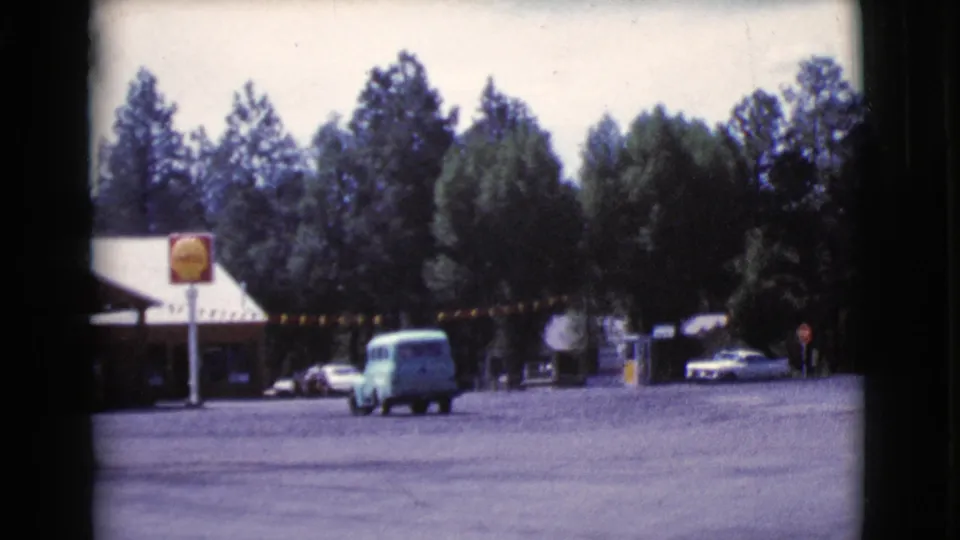 1964 Cottonwood, Arizona roadside gas station viewed across a paved lot as a light blue van drives past a yellow sign and low building.