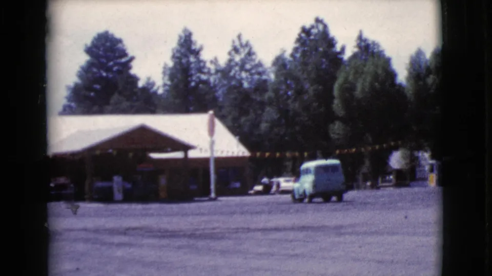 The van continues across the Cottonwood roadside lot, with the service building, parked cars, bunting, and tall trees in the background.