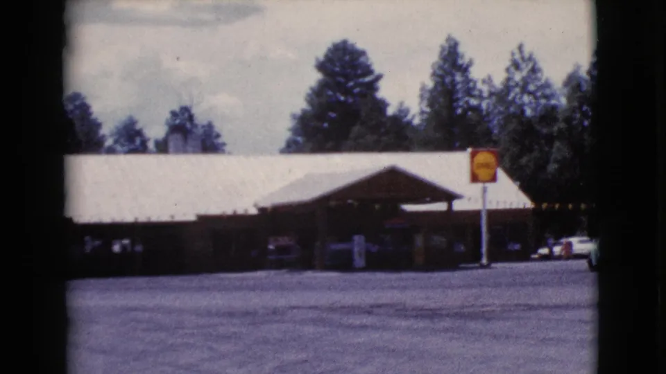 A broader view of the Cottonwood gas station shows the white roof, yellow sign, parked cars, open pavement, and surrounding trees.