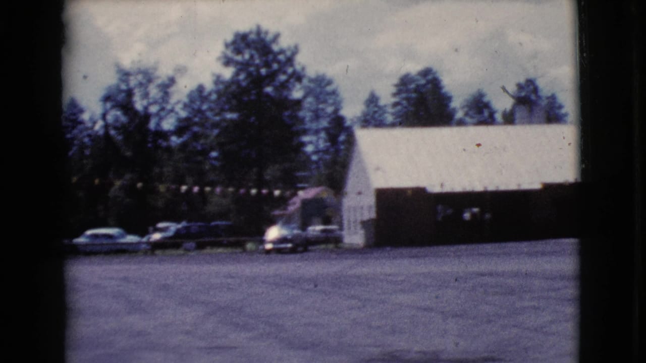 Motel parking lot with 1960s cars, a white gabled building, bunting, and trees.