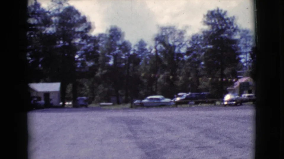 Wide 8mm view of a Cottonwood motel parking area with cars, trees, a small outbuilding, and the motel sign near the right edge.