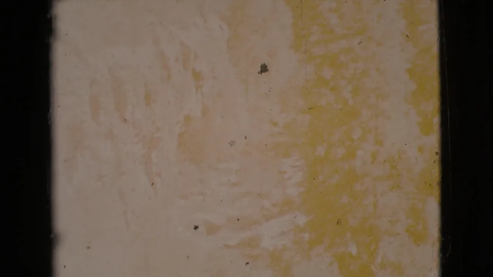 Washed-out 1964 8mm frame of a rocky surface with yellow staining, dust specks, and heavy dark edge framing.