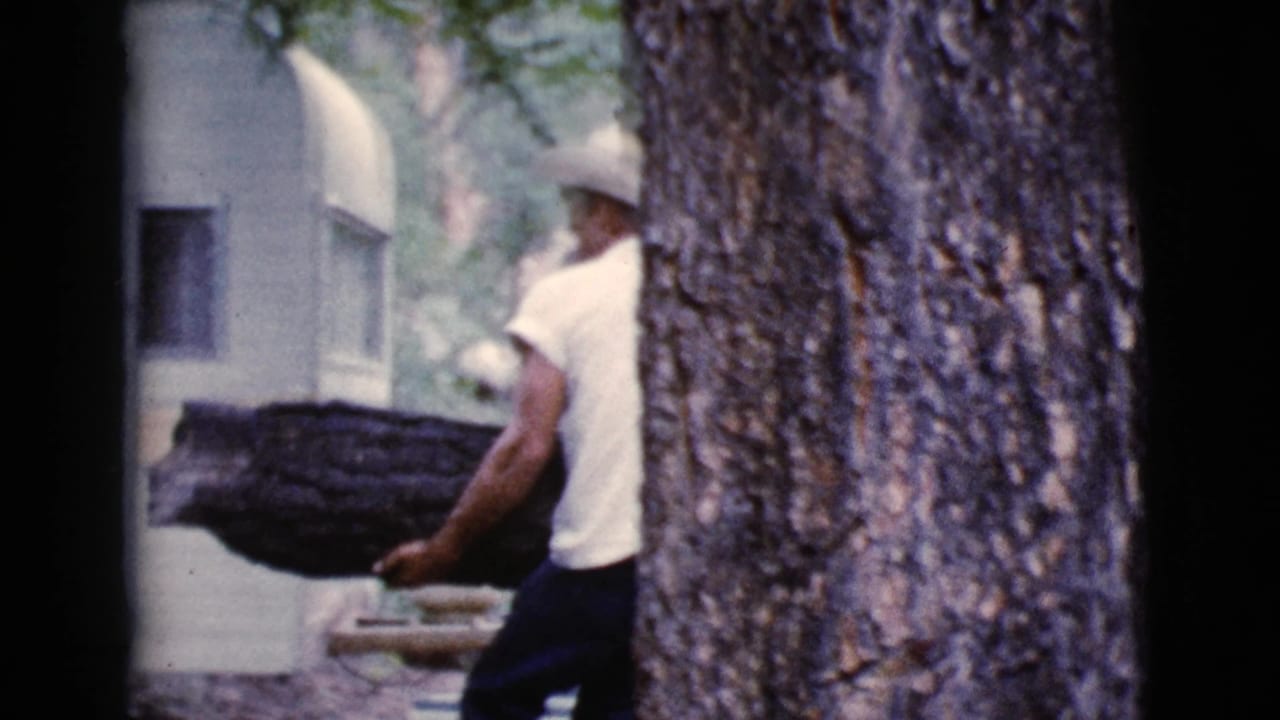 Man in a cowboy hat carries a large log beside a white trailer and tree.