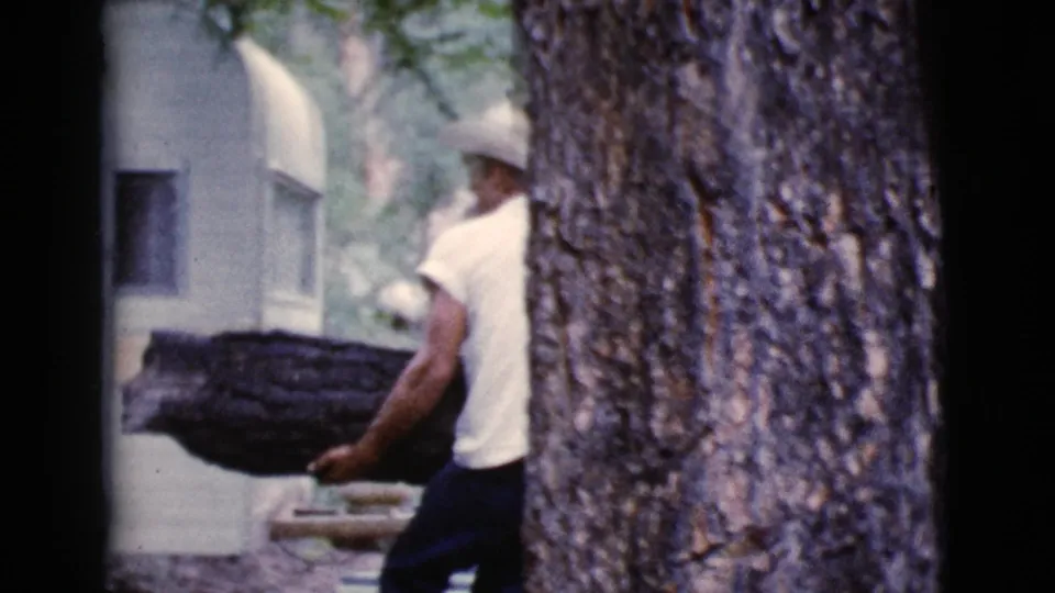 Man in a white shirt and cowboy hat carries a large firewood log near a white trailer in Cottonwood, Arizona, 1964.