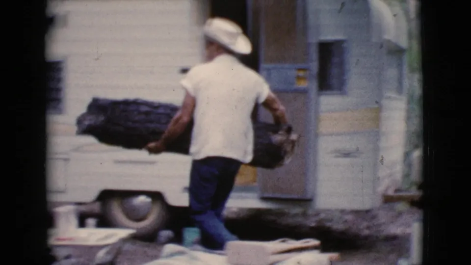 Rear view of a man in a cowboy hat carrying firewood past a white trailer, with 8mm grain and soft color visible.