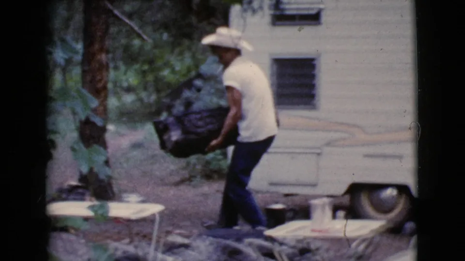 Man carries a large log toward a wooded area beside a white trailer and small table in 1964 Cottonwood, Arizona footage.
