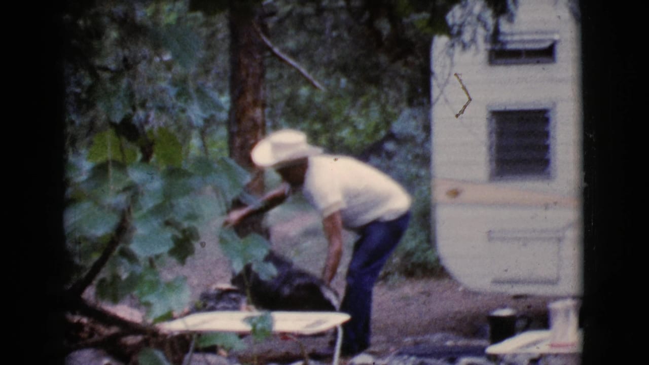 Man in a white cowboy hat bends near a small table at a wooded campsite.