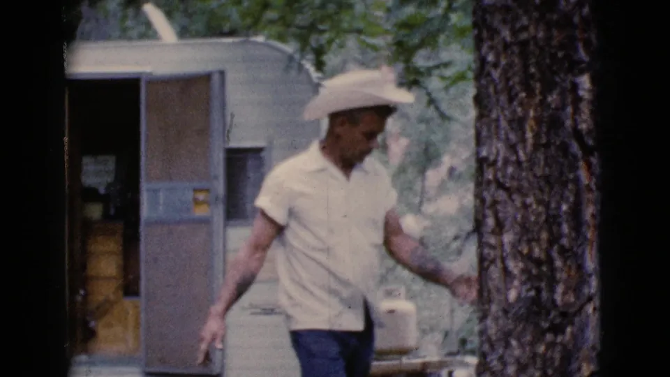 A man in a cowboy hat passes the open camper trailer door, with a large tree trunk in the foreground of the campsite scene.