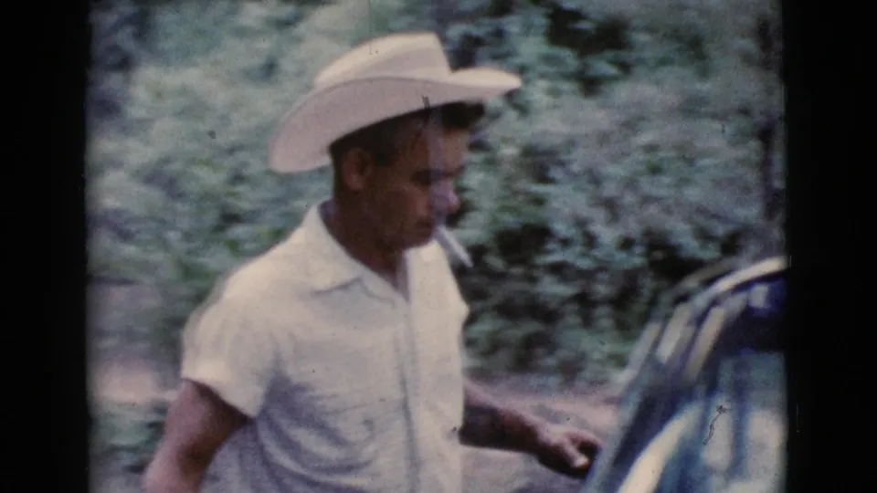 The man walks past a car at the wooded Cottonwood campsite, holding a small object as the 8mm frame softens in motion.
