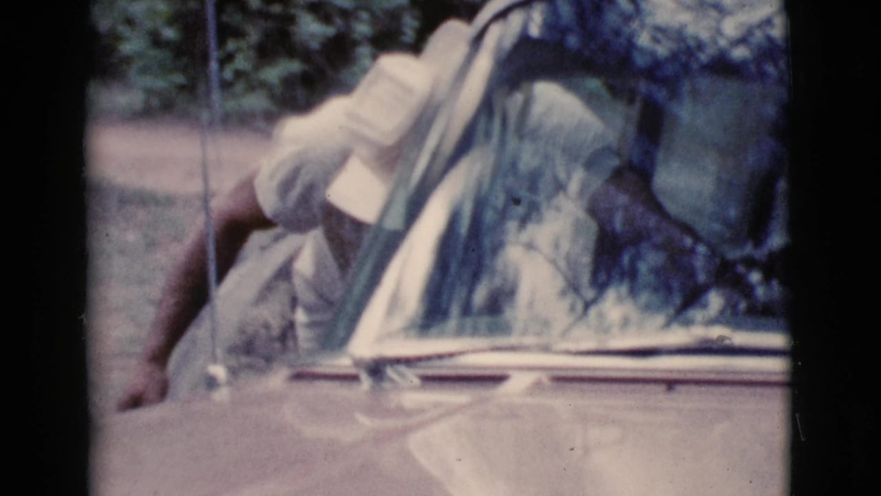 Man in a white hat leans beside a pale vintage car windshield.