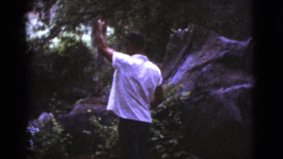 The man is seen from behind in a white shirt and hat, raising one arm beside rocks and foliage in the Cottonwood outdoor setting.