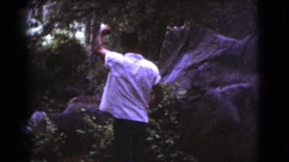 In the final frame, the man remains near a broad rock surface and trees, his arm raised within the shaded 8mm home-movie scene.