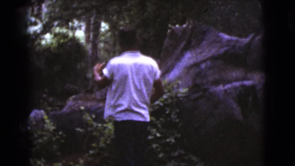 In supplied 1964 Cottonwood, Arizona footage, a man in a white shirt moves through dense foliage near large rocks.