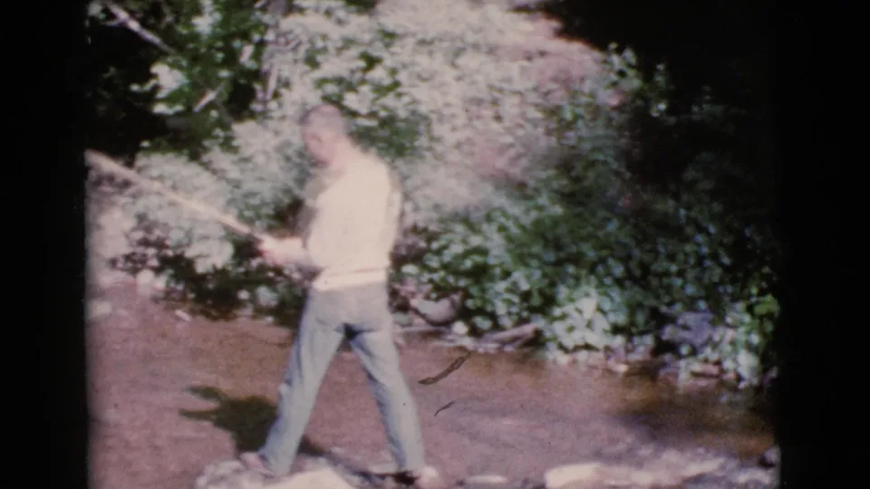 A man holds a fishing pole beside the shallow Cottonwood, Arizona stream, with rocks and dense green foliage in the background.