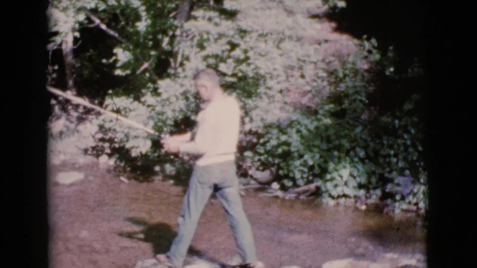 In 1964 Cottonwood, Arizona footage, a man in jeans stands in a shallow stream while holding a fly fishing rod near dense greenery.