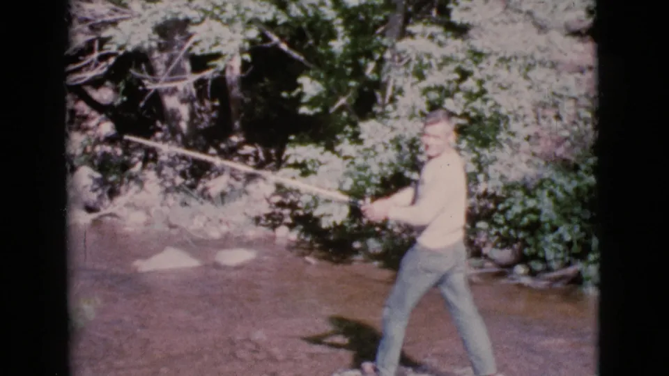 A man in a light shirt and jeans stands in the Cottonwood stream footage, holding his fly rod toward the shaded bank and rocks.