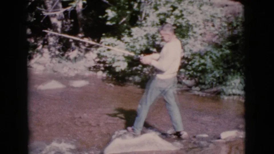 In Cottonwood, Arizona, a man in casual 1960s clothing stands in a shallow stream with a fishing rod under bright sun.