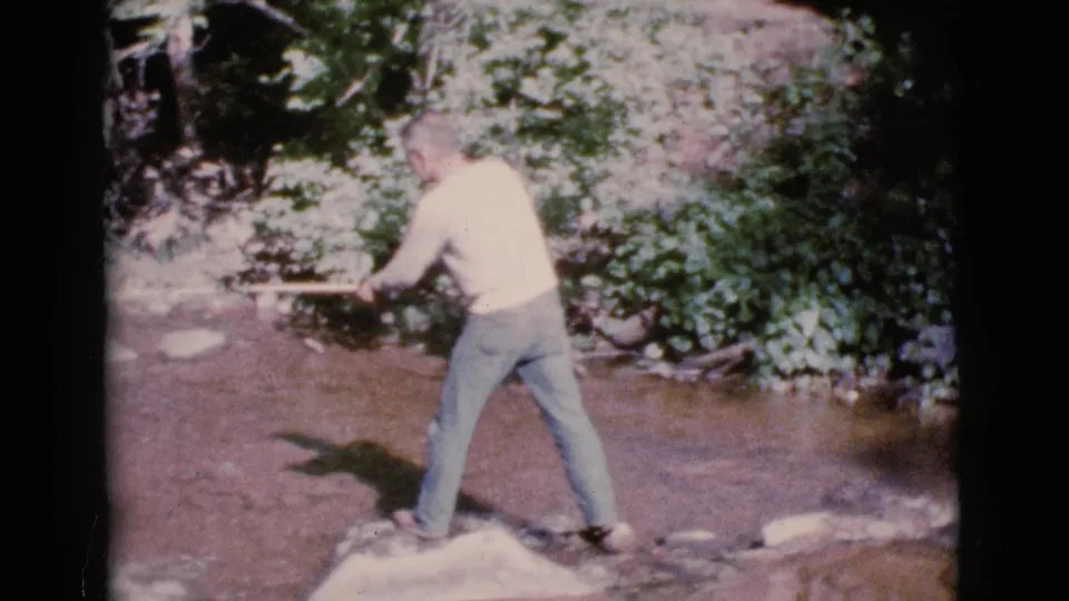 Adult man fishes from a rock in a shallow creek in 1964 Cottonwood, Arizona home-movie footage.
