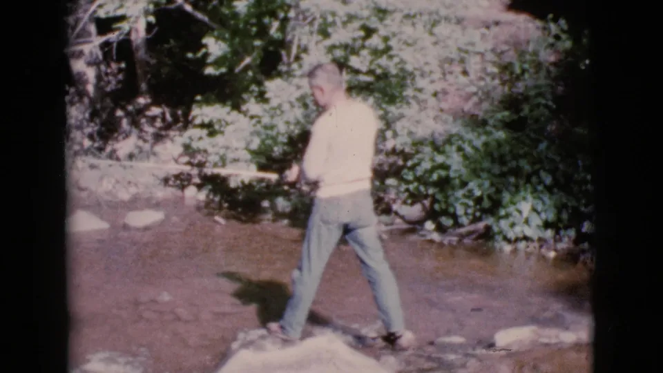 In Cottonwood, Arizona, an adult man stands in creek water and holds a fishing pole toward the leafy bank.