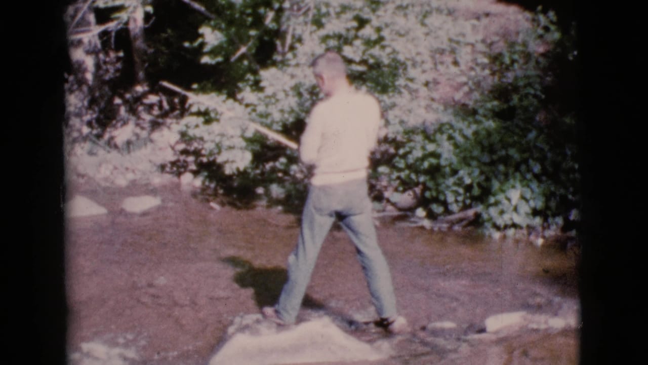 Man in a light sweater and jeans fishing from rocks in a shallow stream.