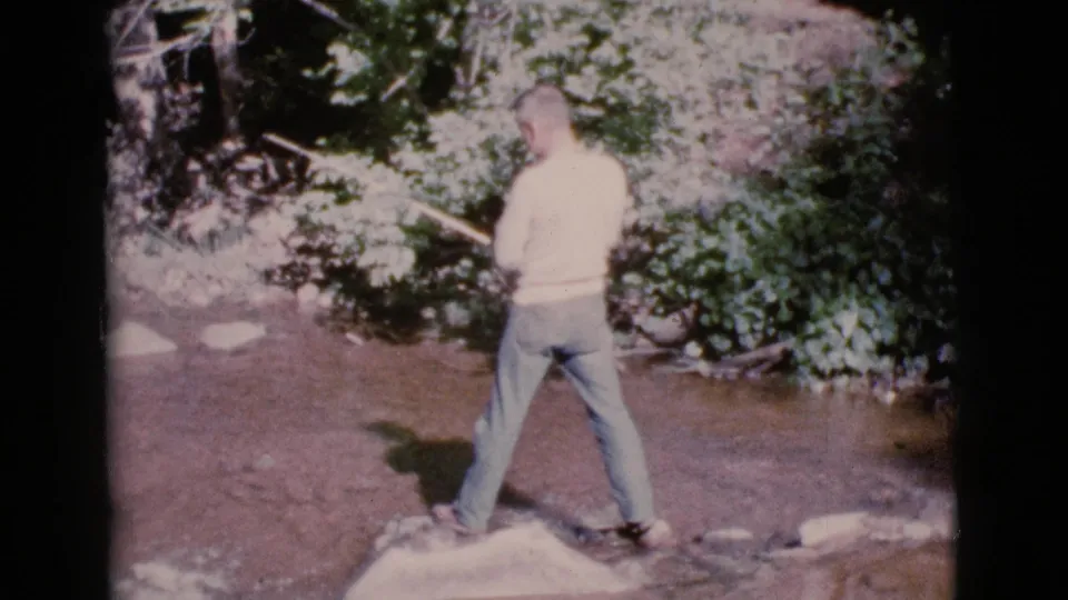 A man fishes with a rod while standing on rocks in a shallow stream near Cottonwood, Arizona, in 1964 home-movie footage.