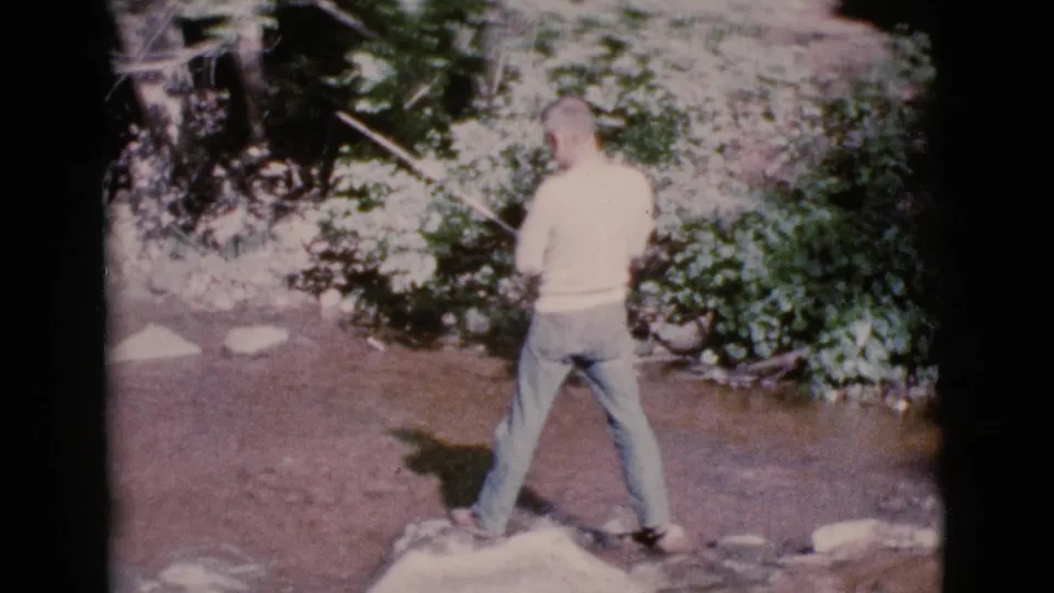 A 1964 home-movie frame shows a man in jeans fishing from rocks in a shallow Cottonwood, Arizona stream.