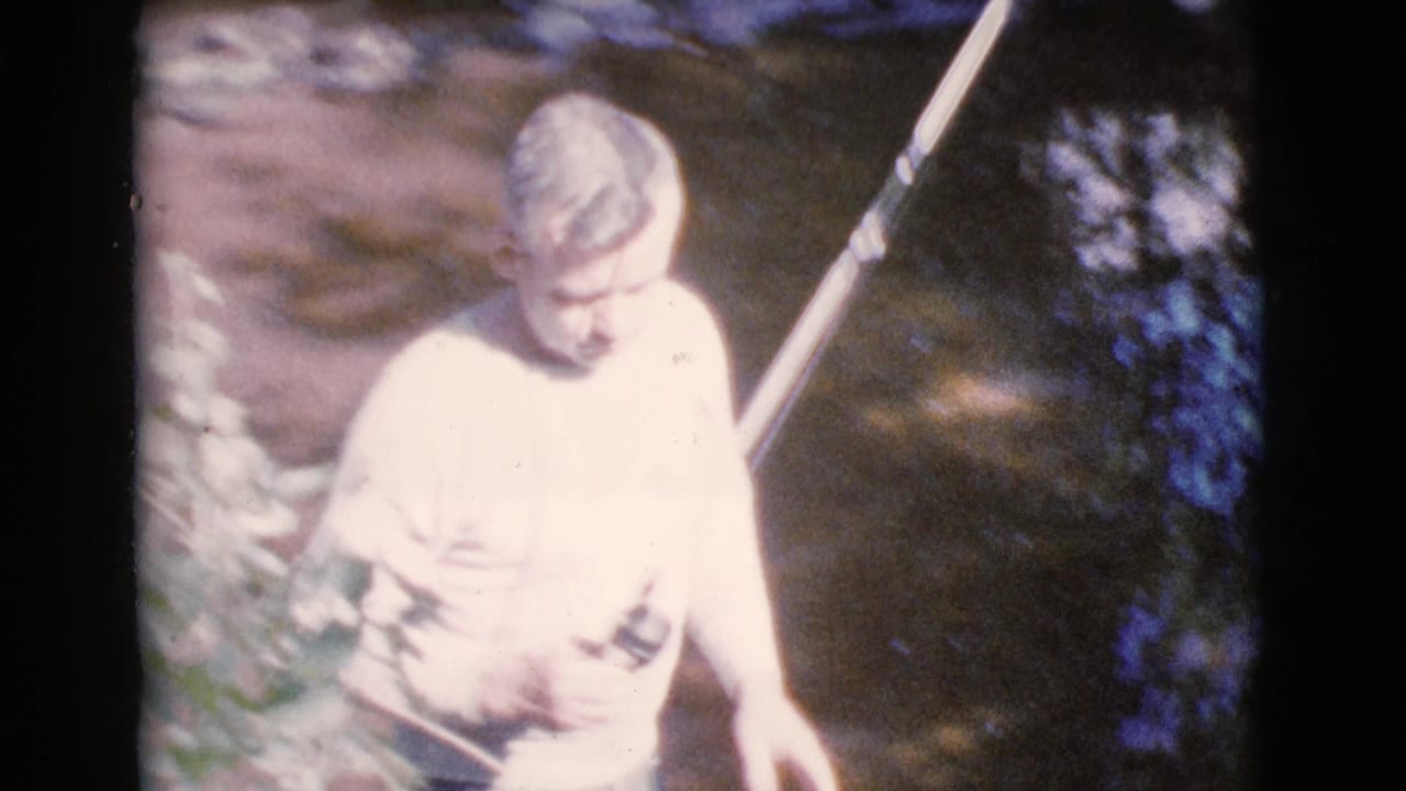 Male figure looks down while holding a fishing rod near streamside plants.