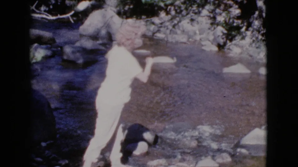In Cottonwood, Arizona, 1964, a woman stands near a shallow rocky stream with a fishing pole while another adult is partly visible behind her.