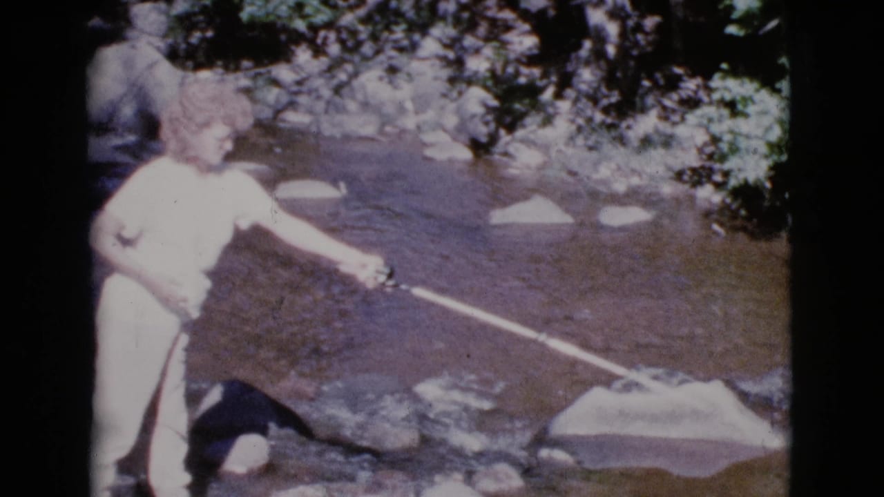 Woman extends a fishing pole over a shallow stream bordered by rocks and trees.