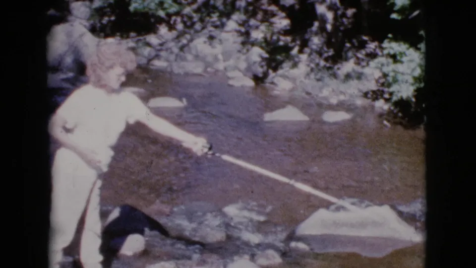 The 1964 Cottonwood home-movie frame shows the woman casting her fishing pole across shallow water with rocks and foliage in the background.