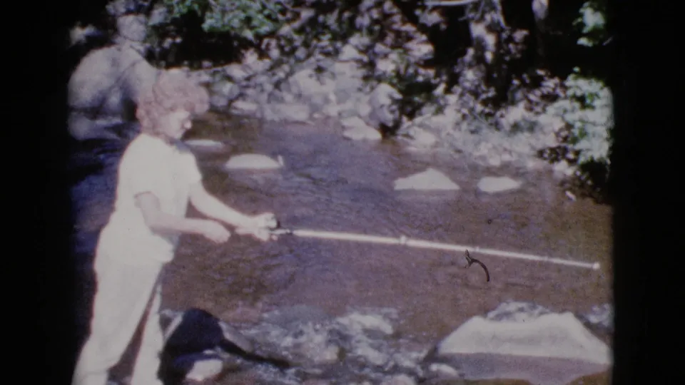 A woman in a light dress fishes at the edge of a shallow rocky stream in Cottonwood, Arizona, in the 1964 home-movie footage.