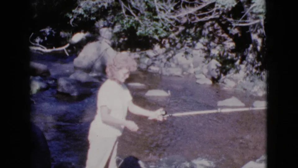 The woman looks toward the water with her fishing pole extended, with a second adult partly visible behind her near the shaded streambank.