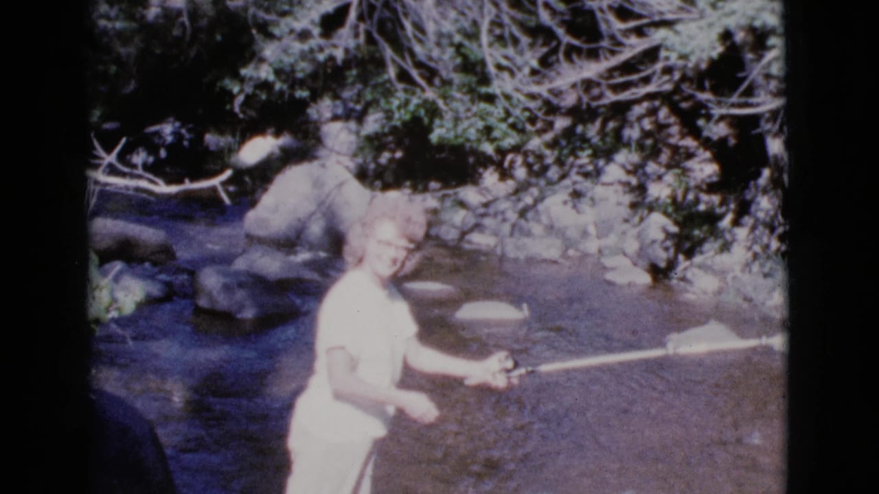 Young woman standing in a shallow stream holding a fishing rod.