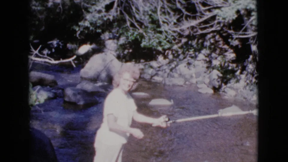 In 1964 Cottonwood, Arizona footage, a young woman in a light dress stands in a shallow stream with a fishing rod.