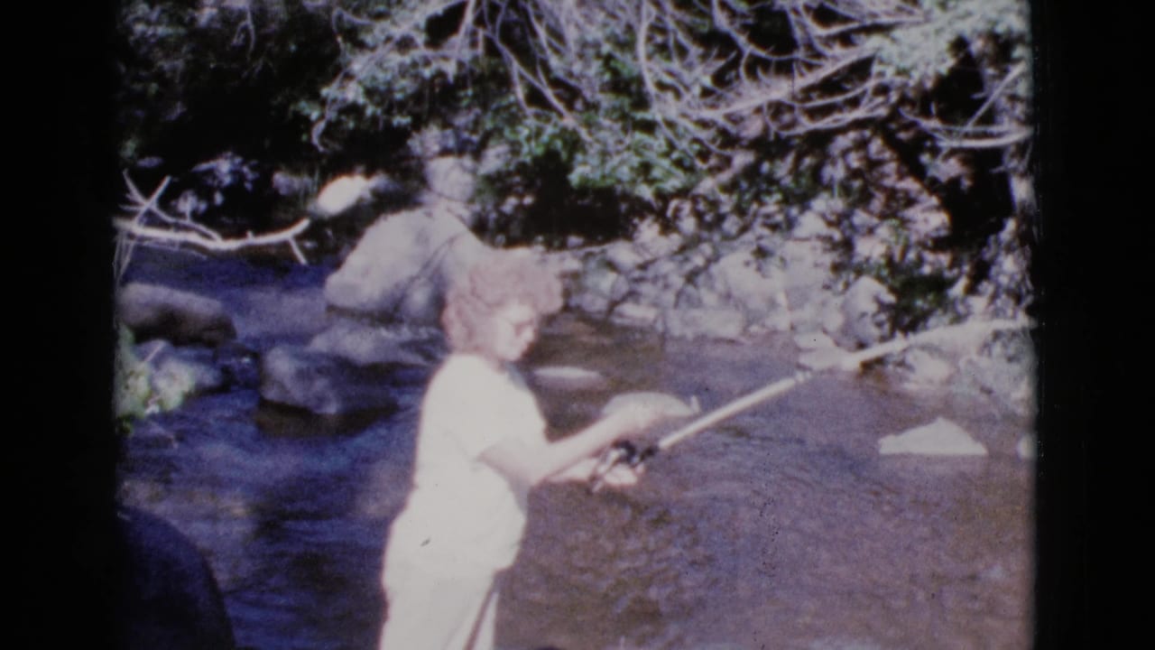 Woman in the stream raises a fishing rod toward the water.