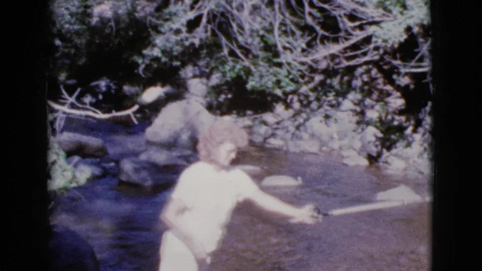 The 1964 home-movie frame shows the woman leaning into her cast with the rod stretched across the stream.