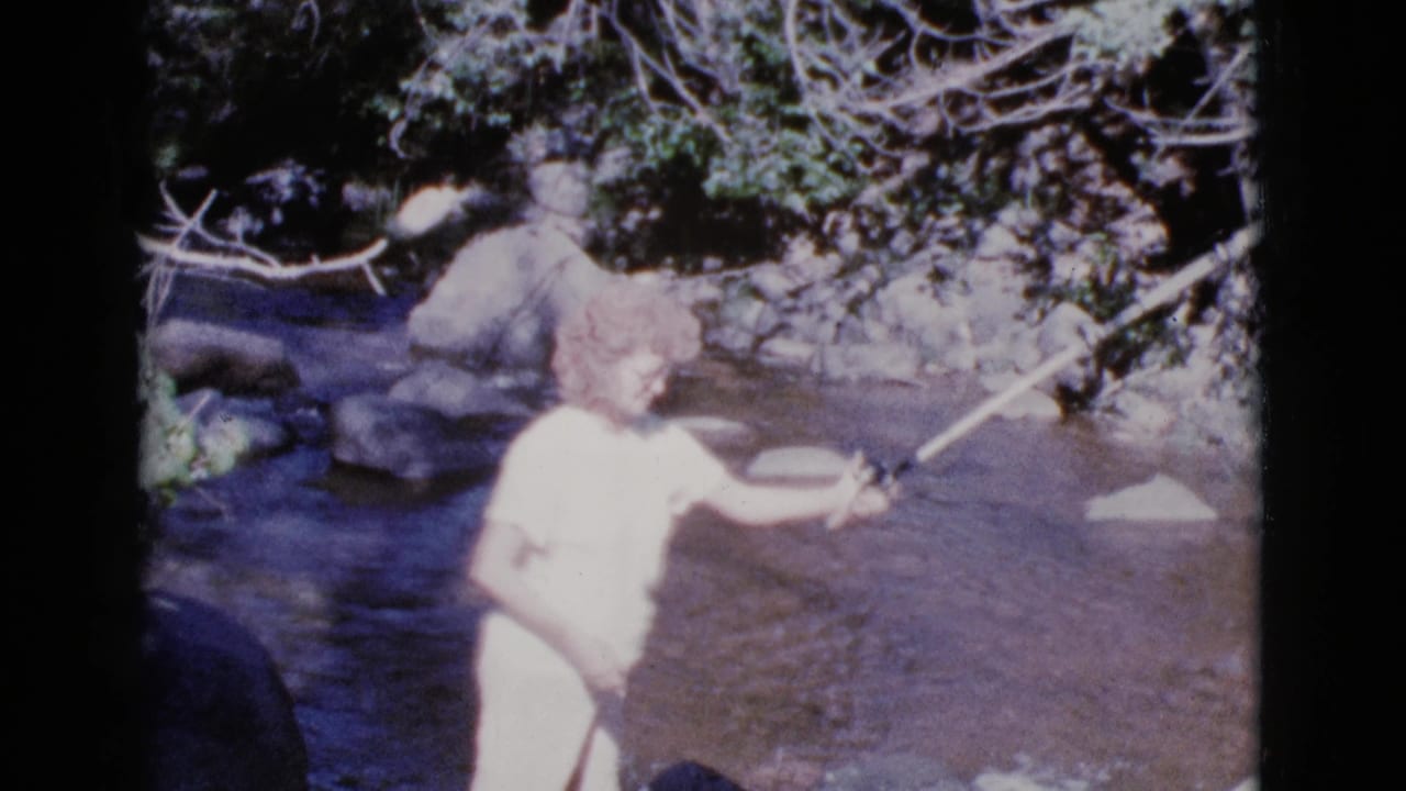 Woman in a white dress stands in a shallow river holding a fishing rod.