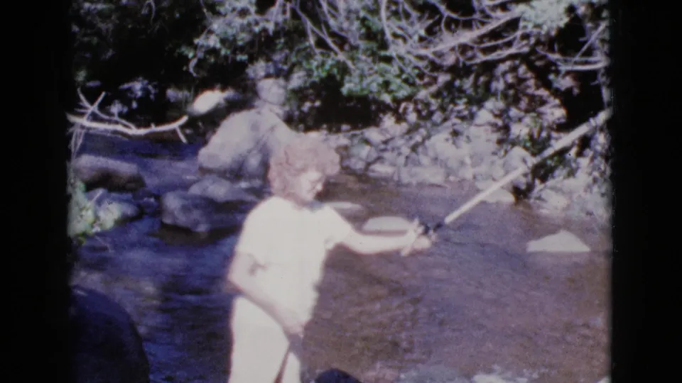 In 1964 Cottonwood, Arizona footage, a woman in a white dress stands in a rocky shallow river while holding a fishing rod.